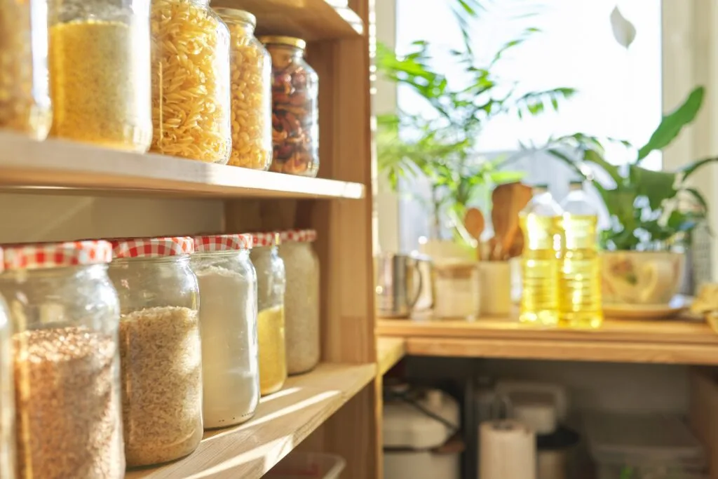 Une étagère de cuisine en bois bien organisée, contenant des bocaux en verre remplis de pâtes, de céréales et d’autres aliments secs, éclairée par la lumière naturelle d’une fenêtre à côté de plantes vertes.